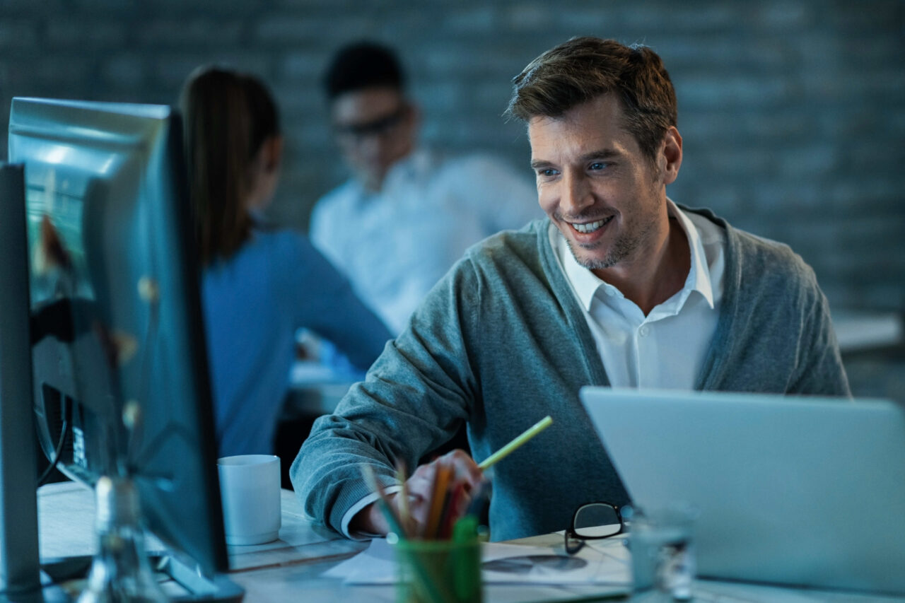 happy-male-entrepreneur-using-desktop-pc-while-working-office-there-are-people-background (1)